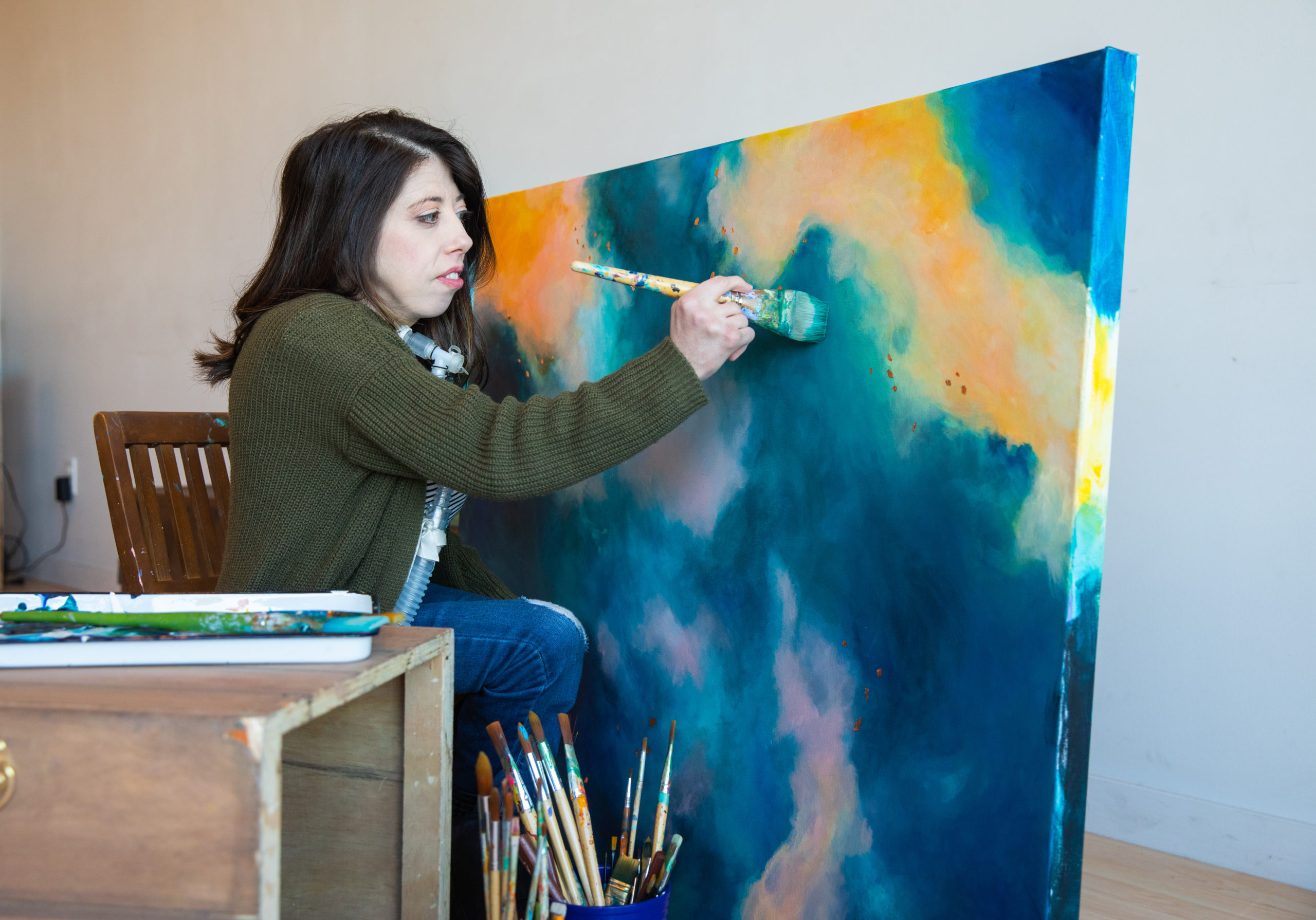 A woman with a disability sitting in a chair while painting on a large canvas